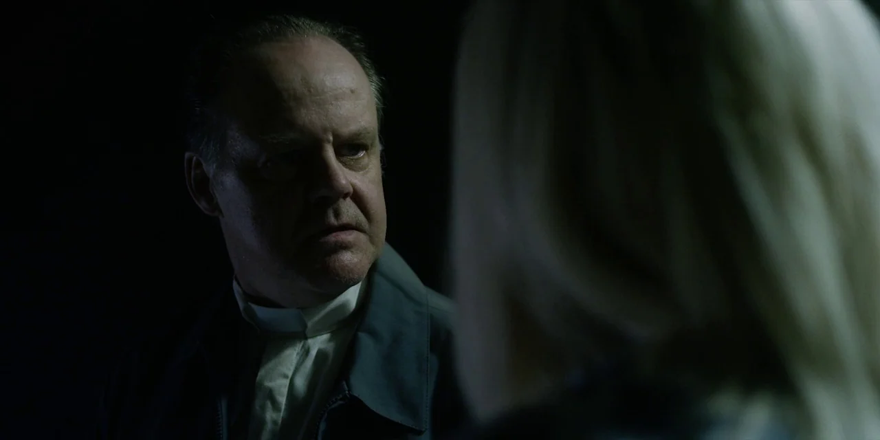 Jakob’s Wife (2021) movie still: medium — A dark, minimally lit interior space serves as the setting for an interaction. A man in clerical collar…