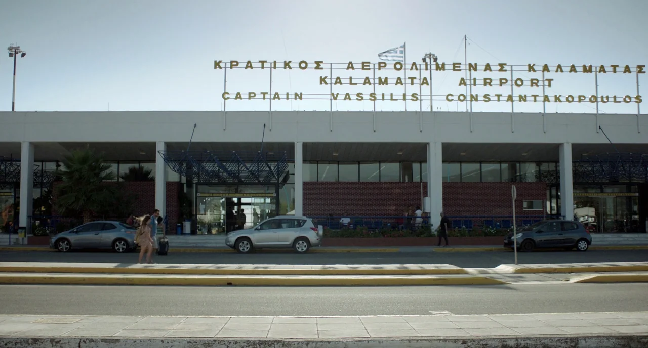 Before Midnight (2013) movie still: wide — A low concrete airport terminal building features prominent Greek and English gold lettering. Several…