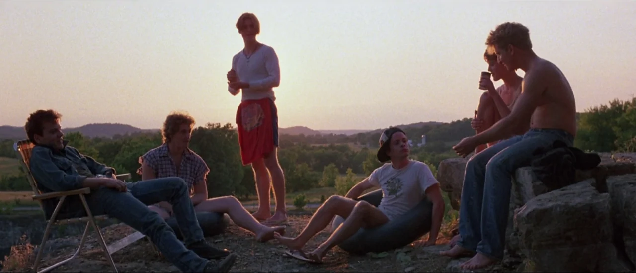 At Close Range (1986) movie still: wide — A group of young people gather on a rocky ridge during a sunset. They sit on folding chairs and rubber…