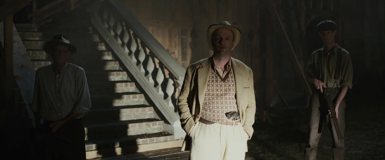 Live By Night (2016) movie still: fullbody — A dark wooden barn interior features a stone staircase with a balustrade in back. Three men stand in a…