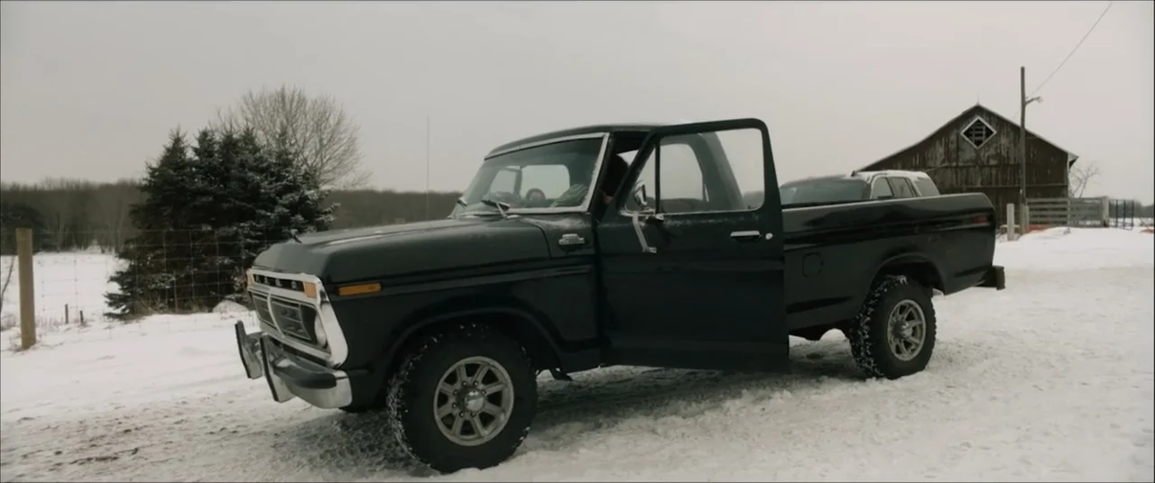 I'll Take Your Dead (2018) movie still: wide — A dark vintage pickup truck sits on a snowy rural road near a barn. A male driver sits behind the…
