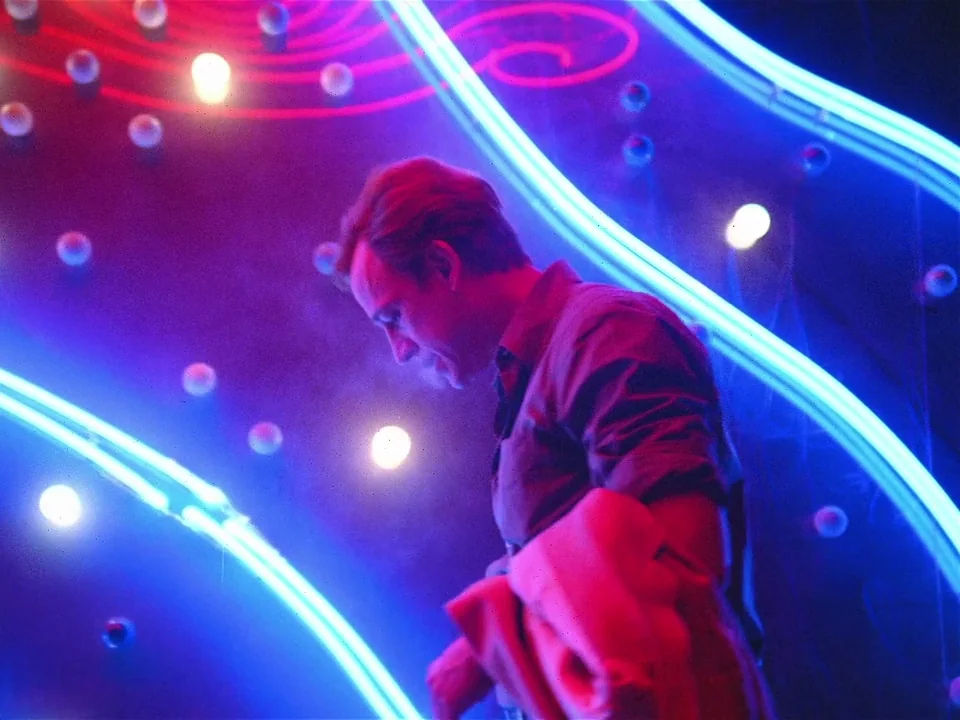 One From The Heart (1982) movie still: medium — A man stands before a wall covered in illuminated neon tubes and exposed bulbs. The person looks…