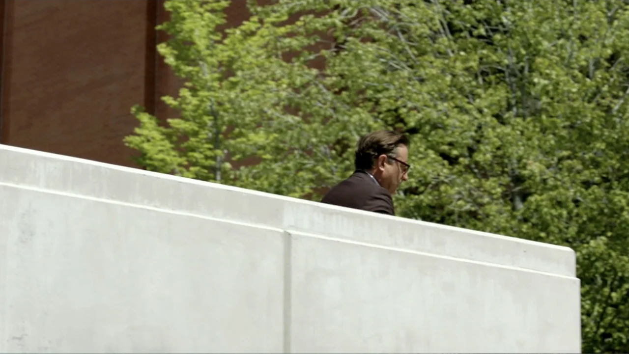 At Middleton (2013) movie still: medium — A man in a suit walks behind a white concrete ledge. Sunlight shines on a green tree in the background.