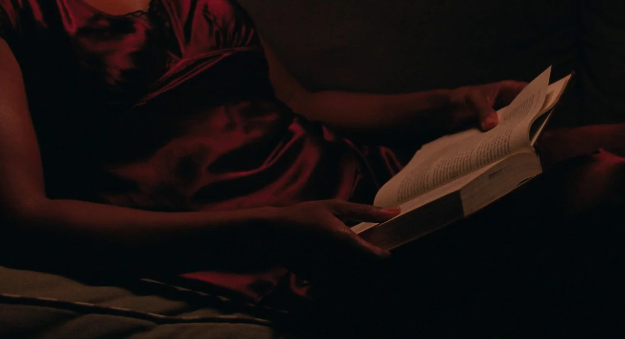Pariah (2011) movie still: medium — A dimly lit scene focuses on a person wearing a silk nightgown holding a book. The individual rests comfortably…