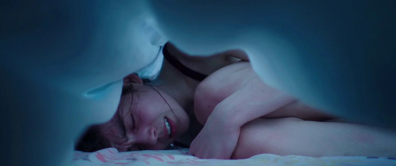 Raw (2016) movie still: closeup — A person lies curled under a heavy blanket on a patterned mattress. The close framing obscures the surrounding…