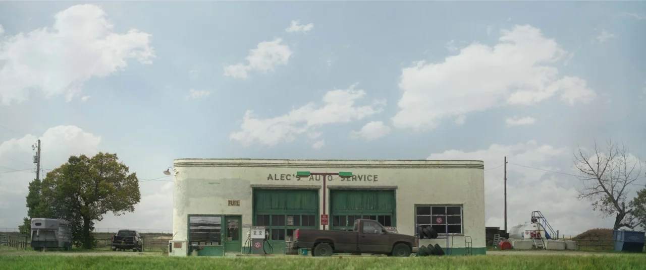 In The Tall Grass (2019) movie still: wide — An aging single-story auto service building features two large green sliding garage doors. A brown…