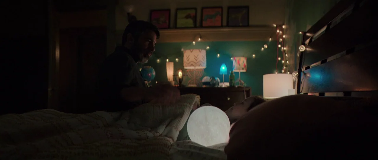 The Boogeyman (2023) movie still: medium — A dimly lit room with teal walls, string lights, and multiple small glowing lamps. An adult man sits on…