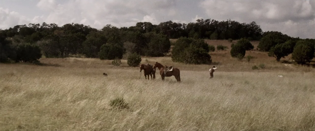 Matar o morir (1984) movie still: wide — A vast, open prairie features tall, dry grass beneath a partially cloudy sky. A person walks toward two…
