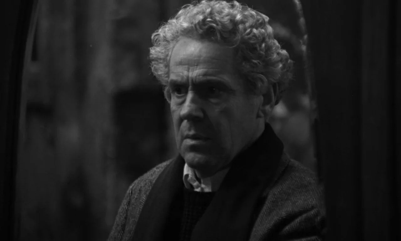 Nostalghia (1983) movie still: closeup — A dark, dimly lit room contains an elderly man with curly, receding grey hair. The man in a dark coat and…