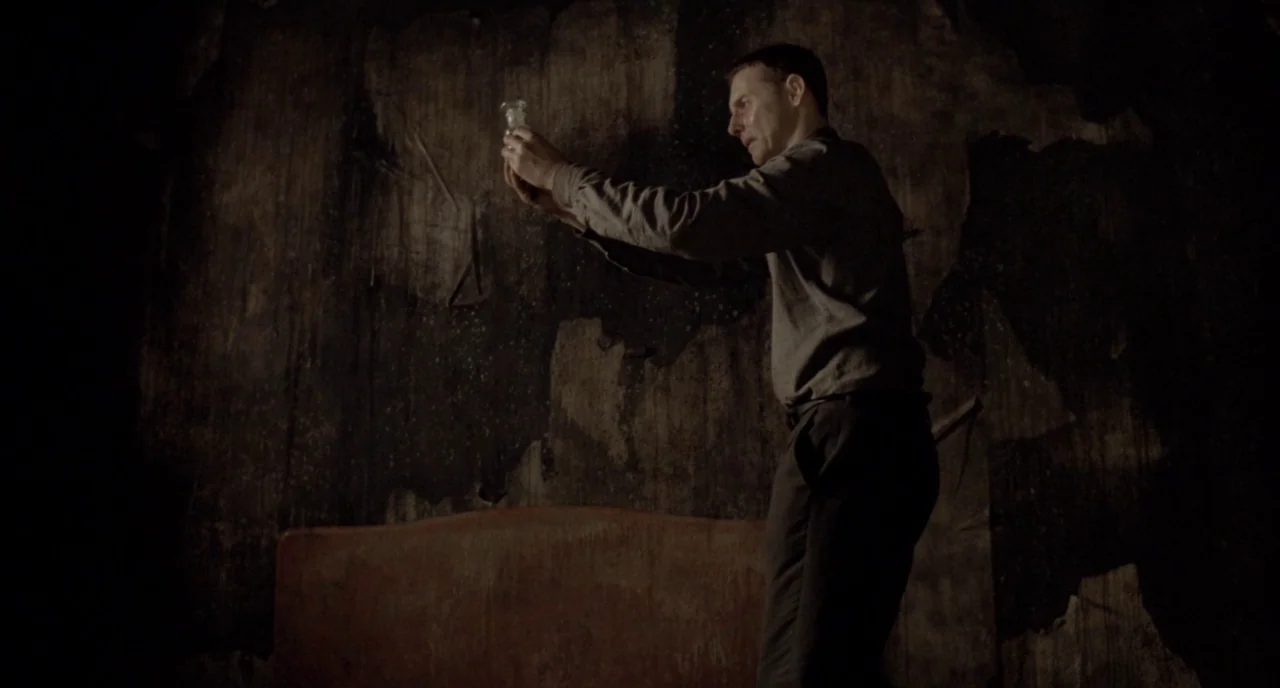 Possum (2018) movie still: medium — A dimly lit room features walls covered in dark, damaged, and peeling wallpaper. A man stands in profile and…