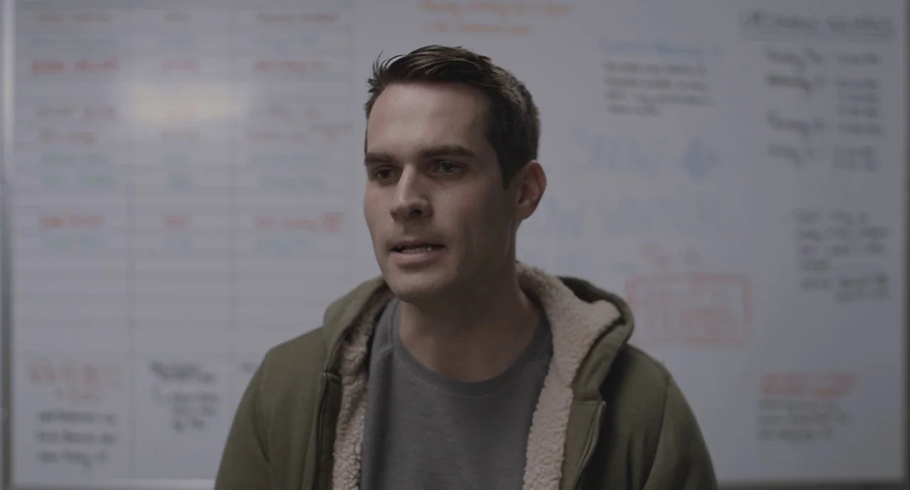 The Wolf of Snow Hollow (2020) movie still: medium — A man stands in an office environment in front of a white board. He speaks to an unseen person…