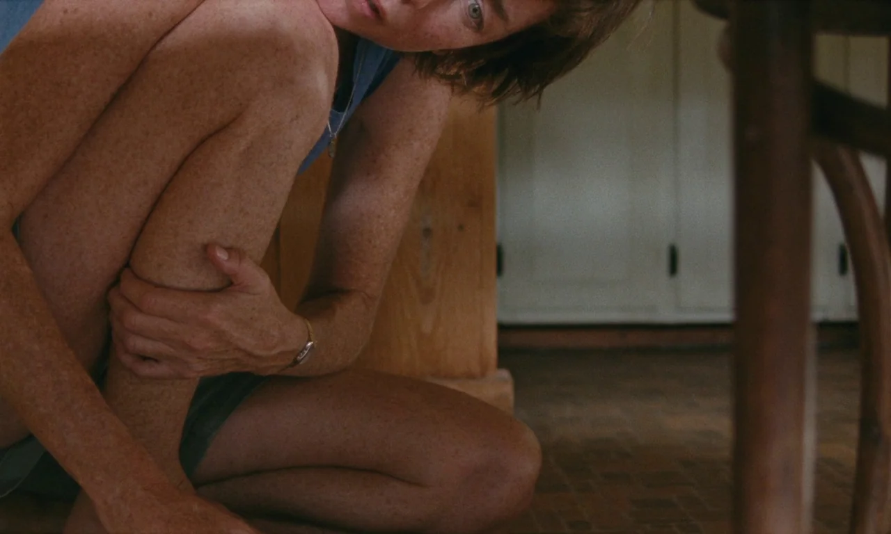 Janet Planet (2023) movie still: medium — A person is crouched on a wood-paneled floor near a large wooden cabinet. The individual clutches their…