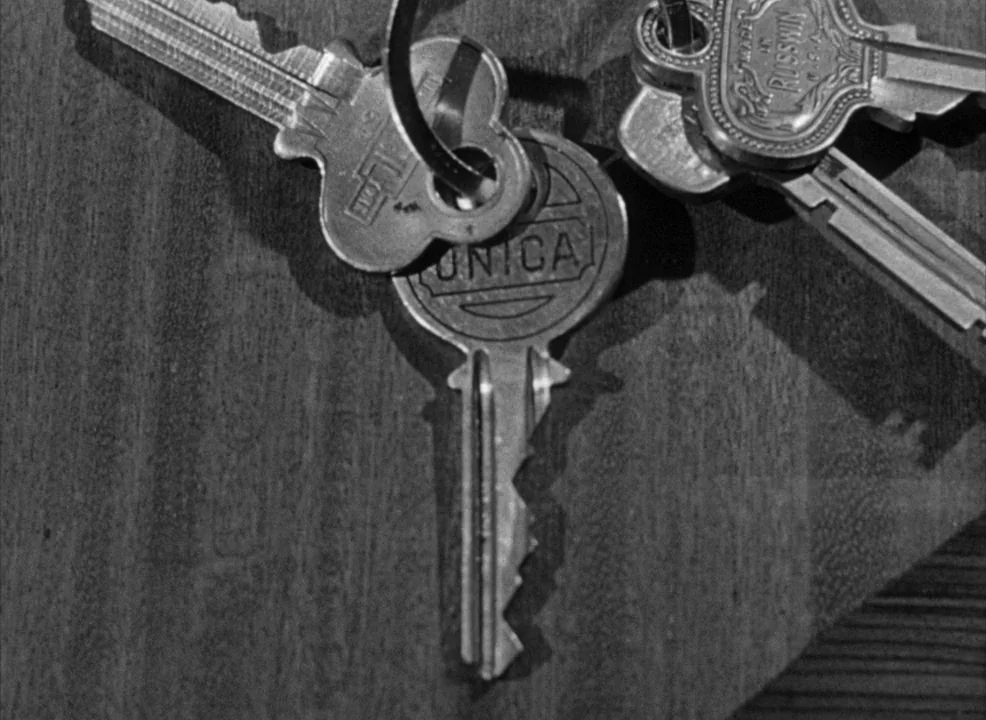 Notorious (1946) movie still: closeup — A close-up view shows three metallic keys on a textured wooden table surface. The keys are arranged in a…