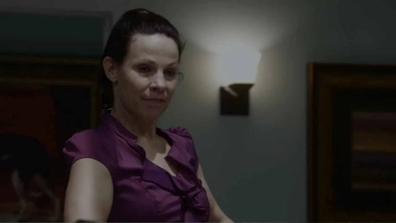 The Messenger (2015) movie still: medium — An interior hallway features muted grey walls and a lit wall sconce. A middle-aged woman in a purple…