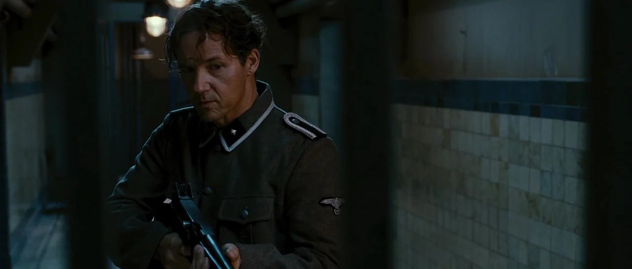 Black Book (2006) movie still: medium — A dimly lit industrial hallway features tiled walls and metal bars in the foreground. A man in historical…