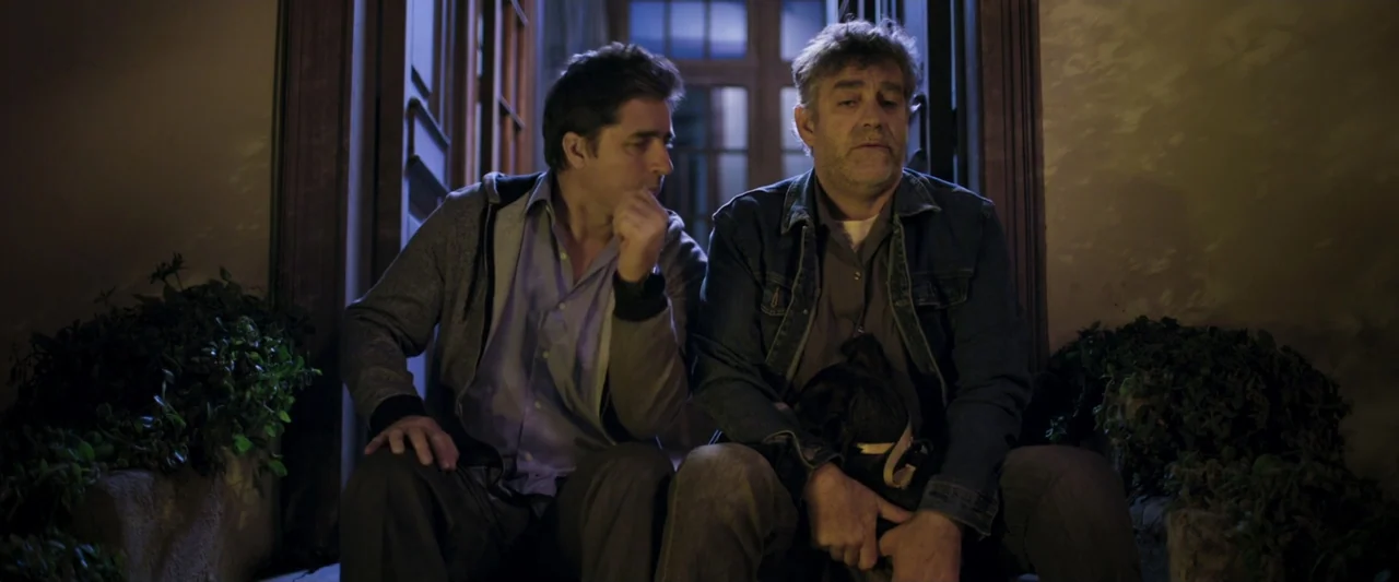 Dog Life (2024) movie still: medium — Two men sit side by side on a step located in a doorway at night. They rest together while one of the men…
