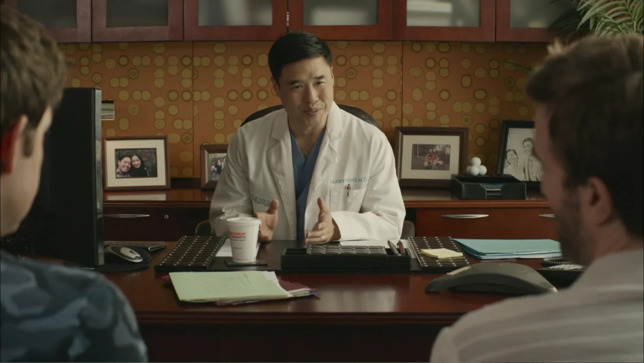 The Hollars (2016) movie still: medium — A professional medical office features a large wood desk with framed family photographs. A male doctor sits…