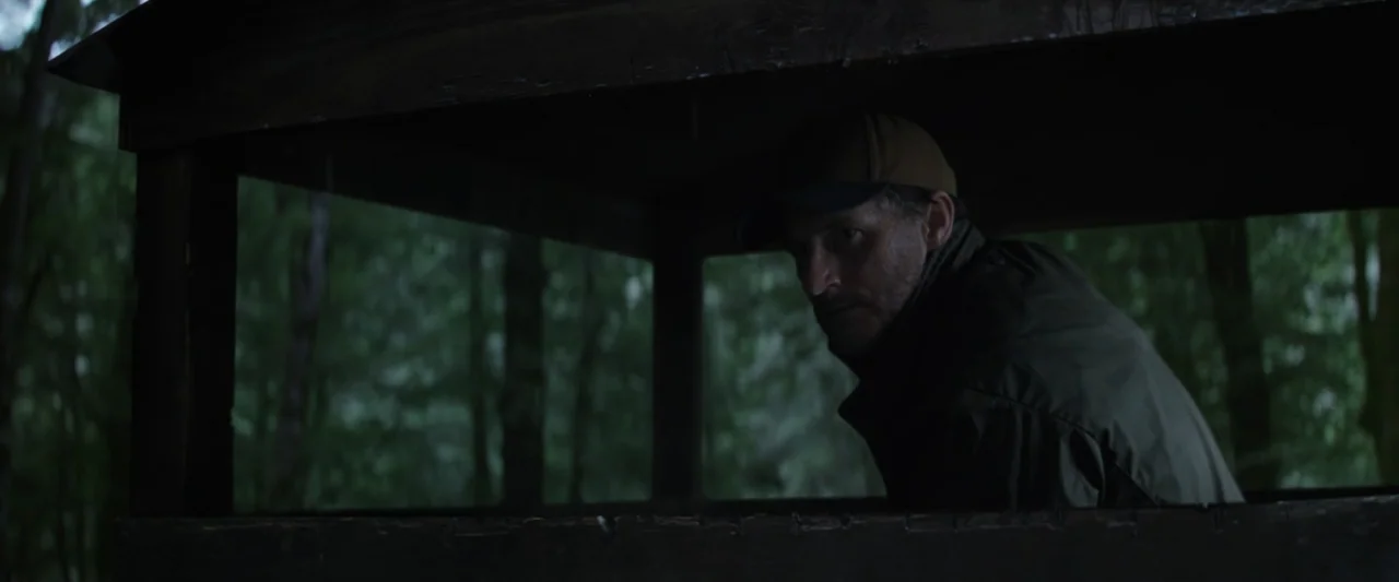 Wolf Man (2025) movie still: medium — A man sits inside a rustic, dark wooden hunting blind surrounded by dense trees. The man glances back over his…
