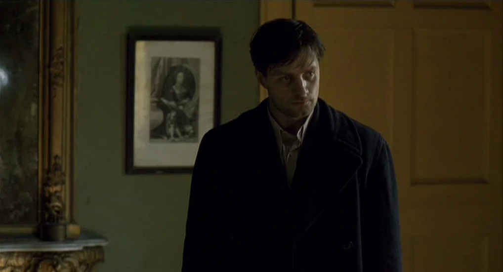 The Wind That Shakes the Barley (2006) movie still: medium — A dimly lit interior space with olive green walls and a framed print. A man in a dark…