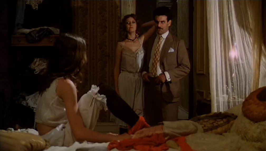 Pretty Baby (1978) movie still: medium — A dimly lit bedroom features ornate patterned wallpaper, sheer curtains, and a cluttered bed. A man in a…