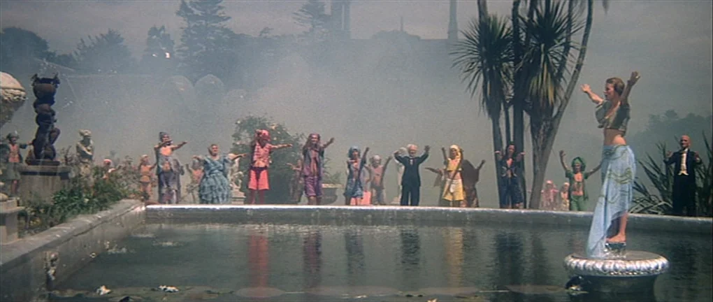Zardoz (1974) movie still: wide — A group of people stands around a fountain in a mist-filled public park area. The subjects stand with arms…