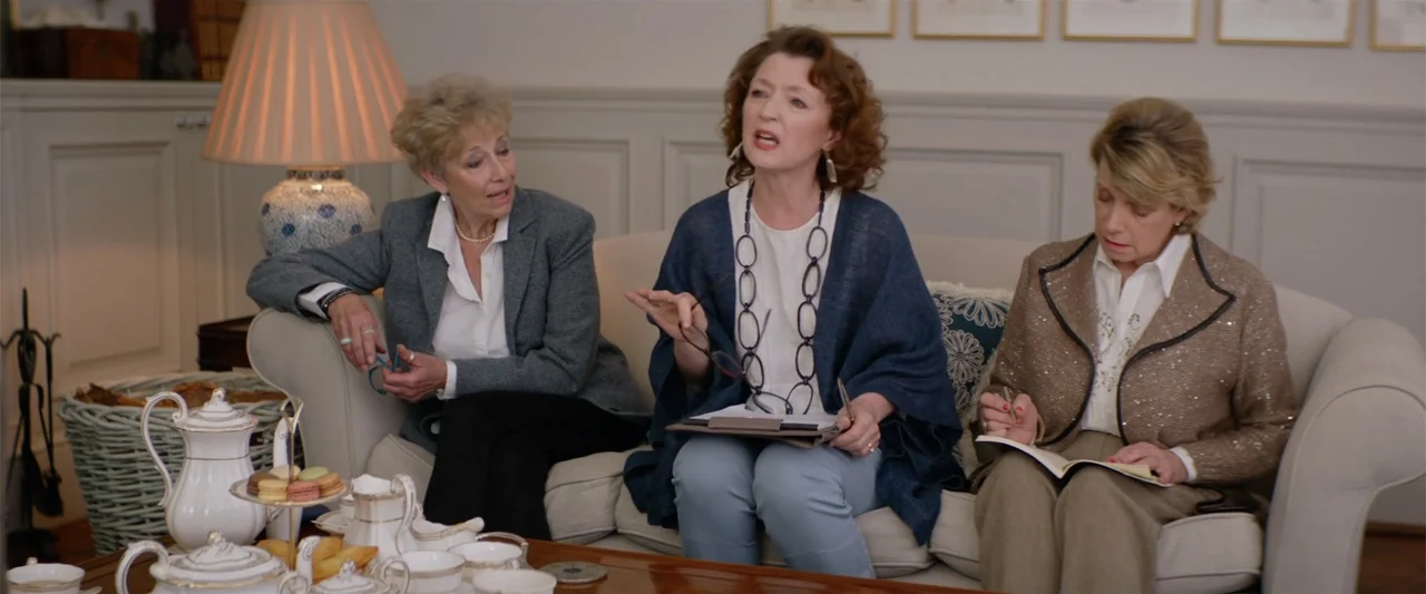 Hampstead (2017) movie still: medium — A sunlit interior features three women sitting on a beige sofa in conversation. A wooden coffee table holds a…