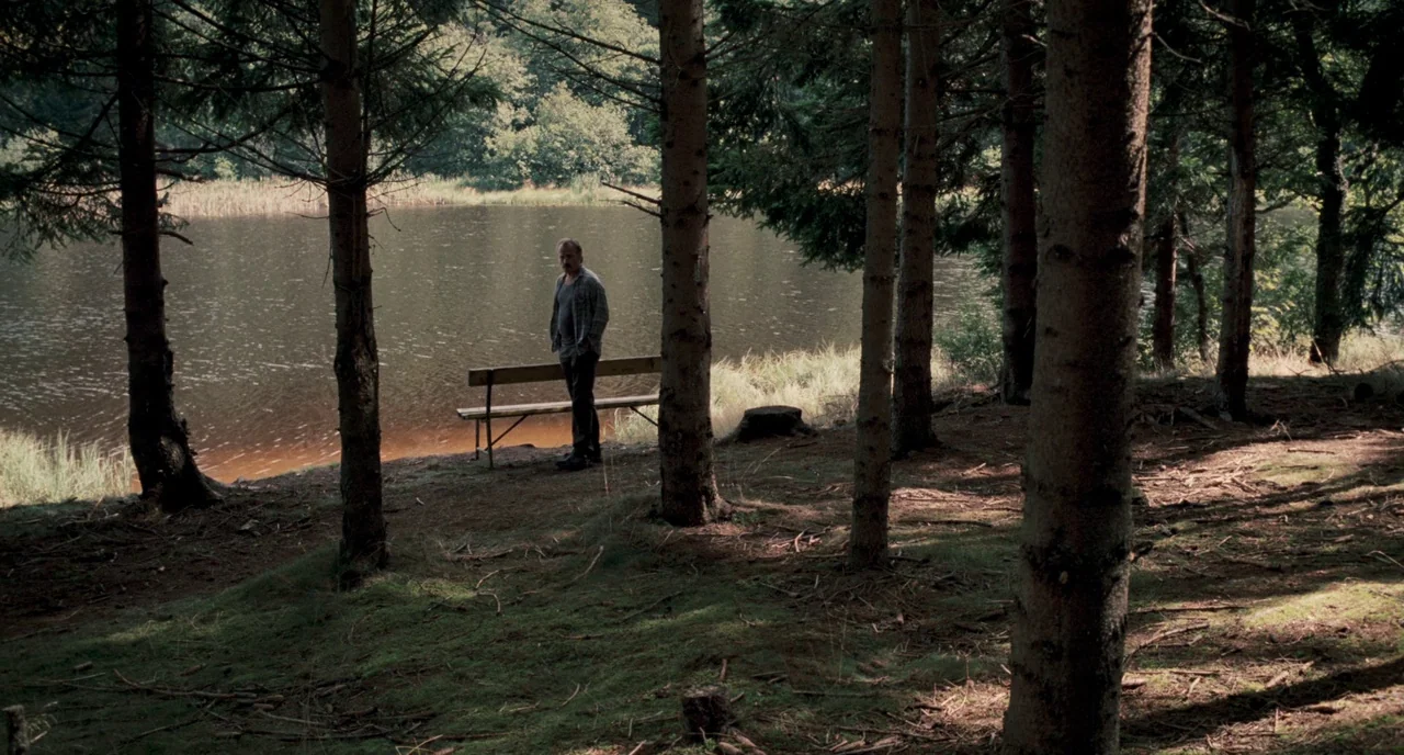 Revanche (2008) movie still: wide — A forested shoreline bordering a still lake with tall pine trees and mossy ground. A middle-aged man stands…