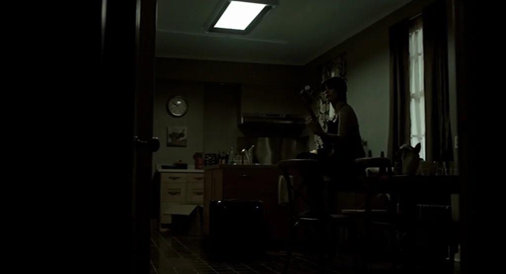Mama (2013) movie still: medium — A dim room features a central wooden island and a dark floor. A woman sits on a chair in the middle of the room…