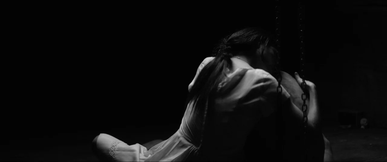 The Eyes of My Mother (2016) movie still: medium — A dark, spartan enclosure features metal chains hanging vertically from the ceiling. A woman in a…
