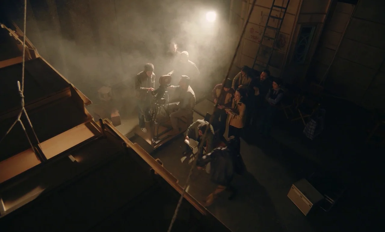 Cobweb (거미집) (2023) movie still: wide — A dimly lit production set interior features thick haze illuminated by a bright overhead light. A film crew…