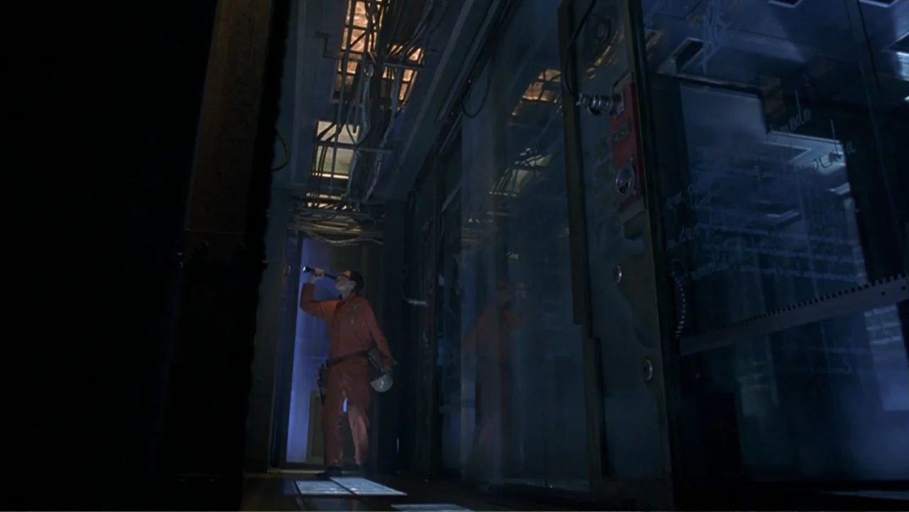13 Ghosts (1960) movie still: wide — An industrial corridor features exposed ceiling conduits and large glass-walled chambers on one side. A lone…