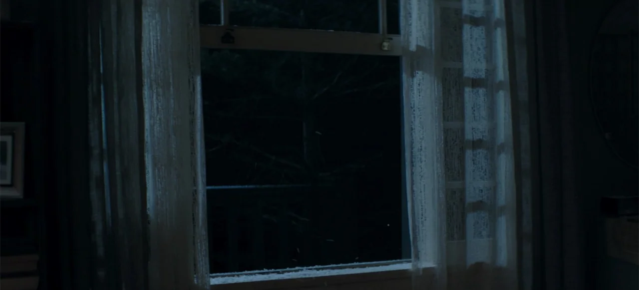 I Still See You (2018) movie still: establishing — A dark bedroom interior features a window framed by thin, light-colored curtains at night. Light…