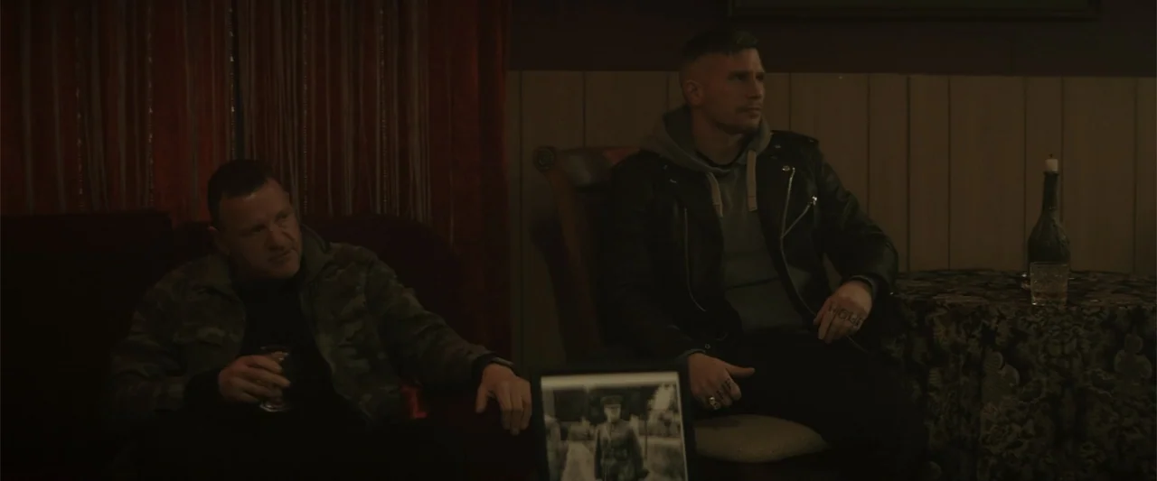 One Ranger (2023) movie still: medium — A dimly lit room features wood paneling and red curtains behind two seated men. The men sit on furniture…