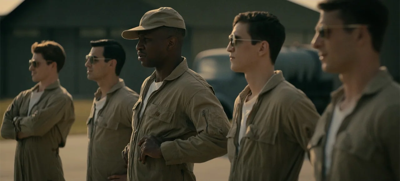 Devotion (2022) movie still: medium — A group of four men stand in a row on a tarmac at sunset. They wear tan flight uniforms and look toward the…