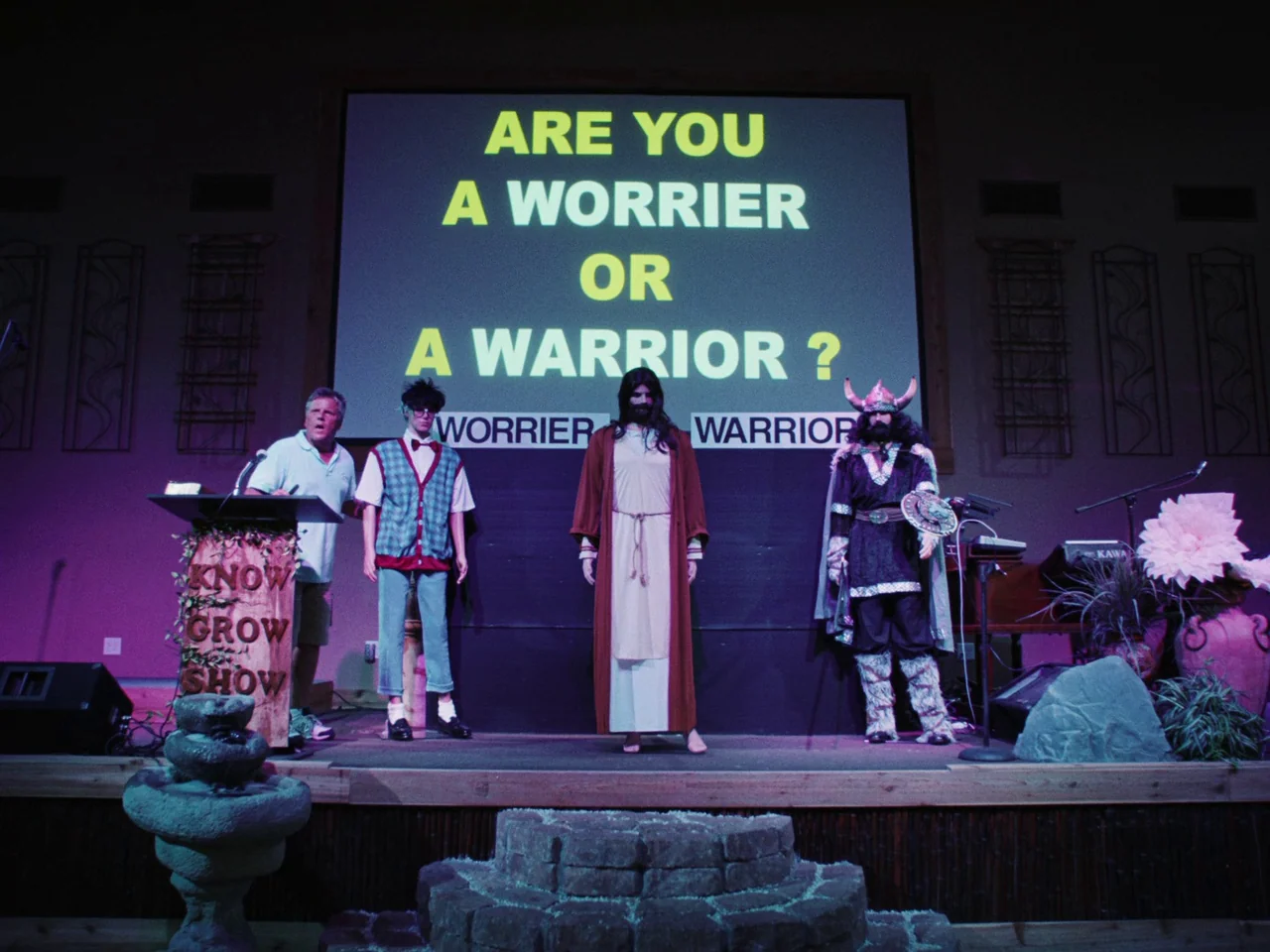 Some Kind Of Heaven (2020) movie still: wide — A wooden stage features four men standing before a large electronic projection screen. The text on…