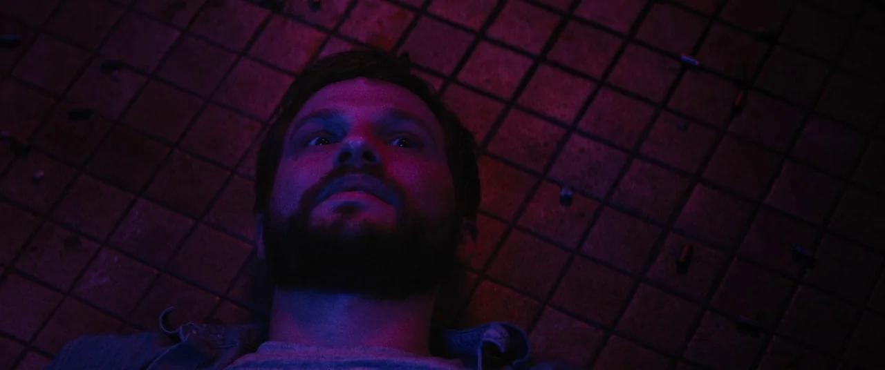 Upgrade (2018) movie still: closeup — A man lies flat on a dark tiled floor with debris scattered nearby. He stares upward with wide eyes under…