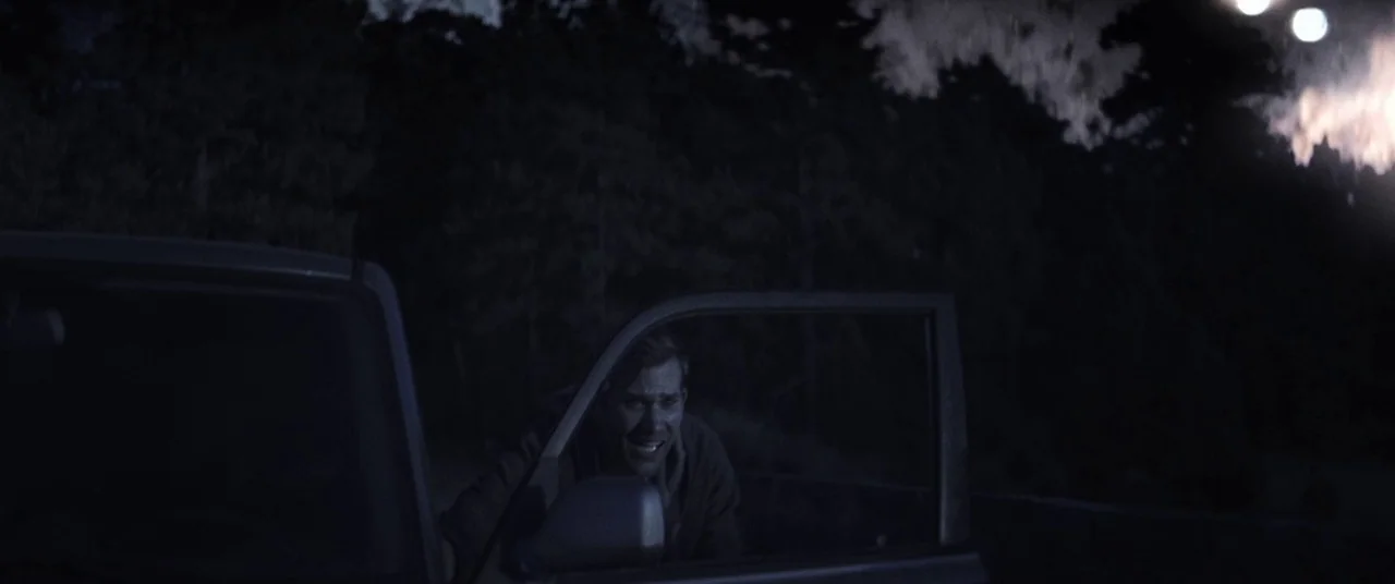 The Endless (2017) movie still: medium — A dark night setting outdoors with trees visible in the background and low light. A man looks out from an…