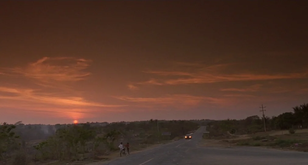 Y Tu Mamá También (2001) movie still: wide — A rural asphalt road stretches into a flat, horizon-filled landscape under a sunset sky. A car with…