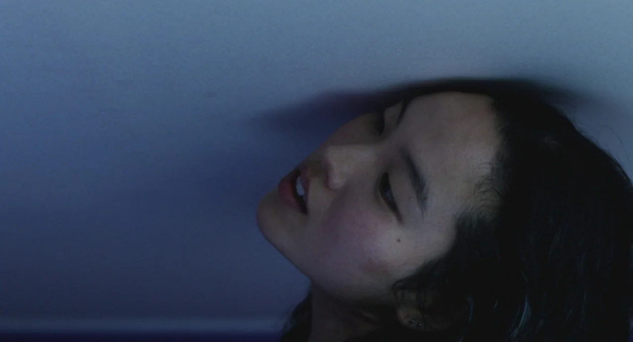 Return To Seoul (2022) movie still: closeup — A close view of a person lying against a wall in a dimly lit room. The individual's face is…
