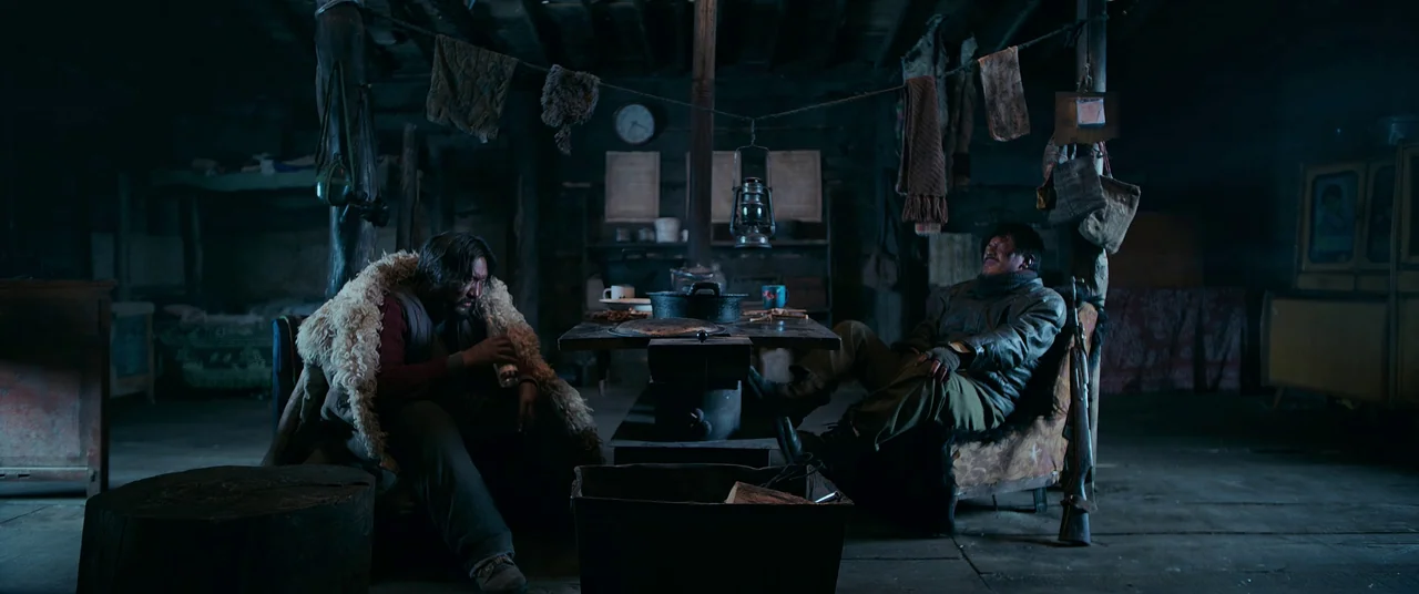 One And Four (2021) movie still: wide — A dark wooden cabin contains a central table, hung laundry, and sparse antique furnishings. Two men rest on…