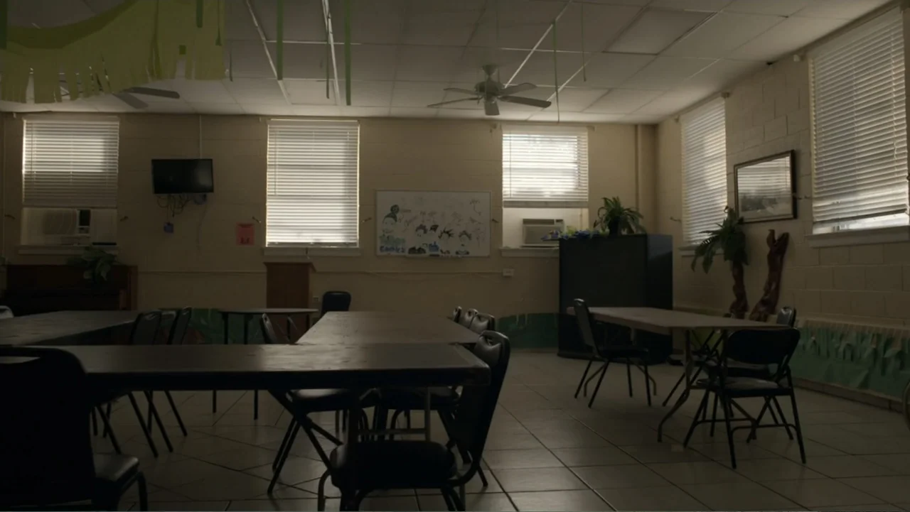 King in the Wilderness (2018) movie still: wide — A quiet community room features rows of folding tables and chairs with beige walls. The space is…