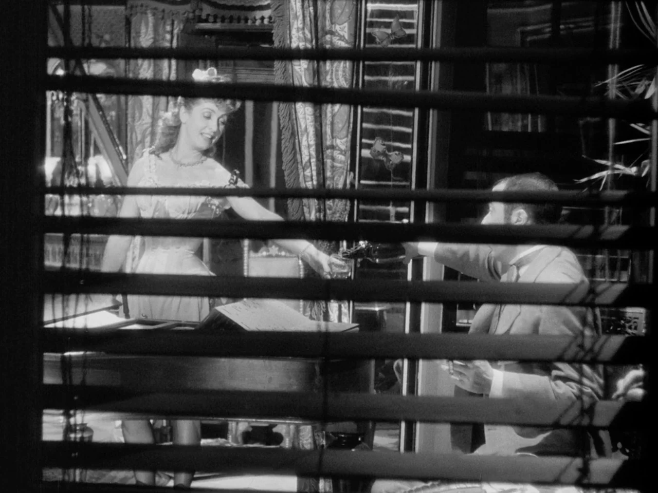 Le Plaisir (1952) movie still: medium — An opulent Victorian parlor is viewed through the slats of wooden Venetian blinds. A woman stands by a piano…