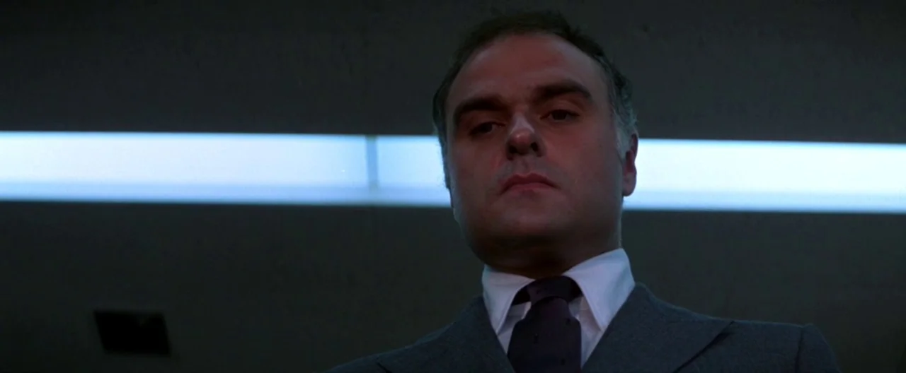 Klute (1971) movie still: closeup — A dimly lit industrial hallway features a single bright horizontal fluorescent light panel. A middle-aged man in…