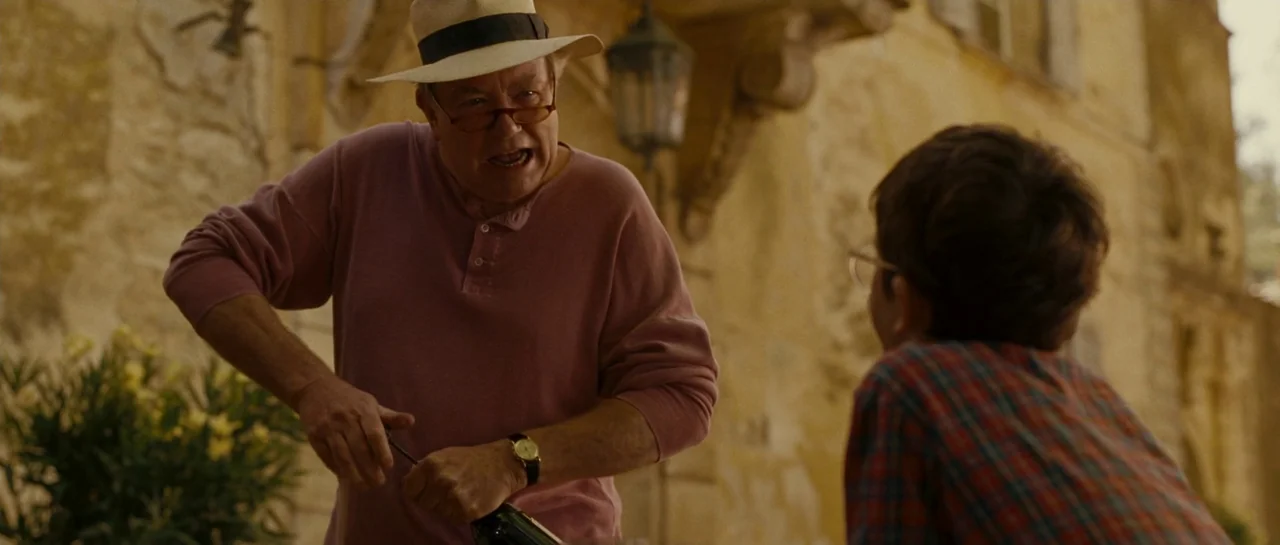 A Good Year (2006) movie still: medium — An older man stands on a sunny stone terrace, pulling a cork from a bottle. A young boy in a plaid shirt is…