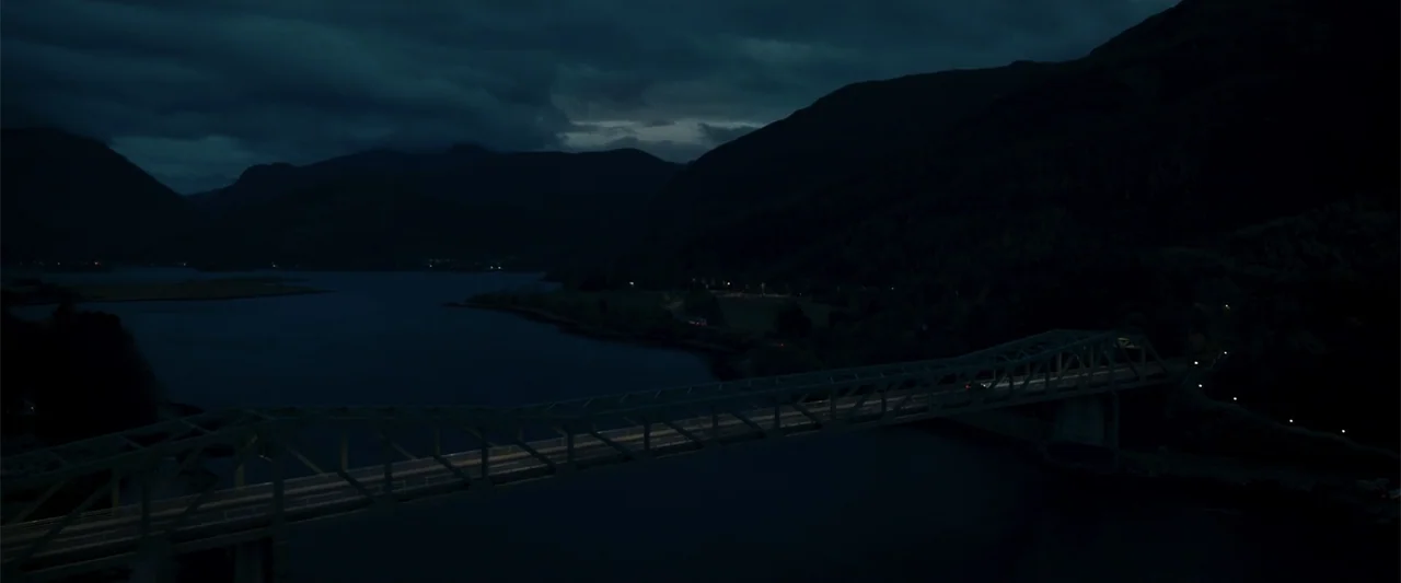 My Son (2021) movie still: establishing — A long bridge crosses a dark river during a cloudy night. Mountains rise in the background with small…