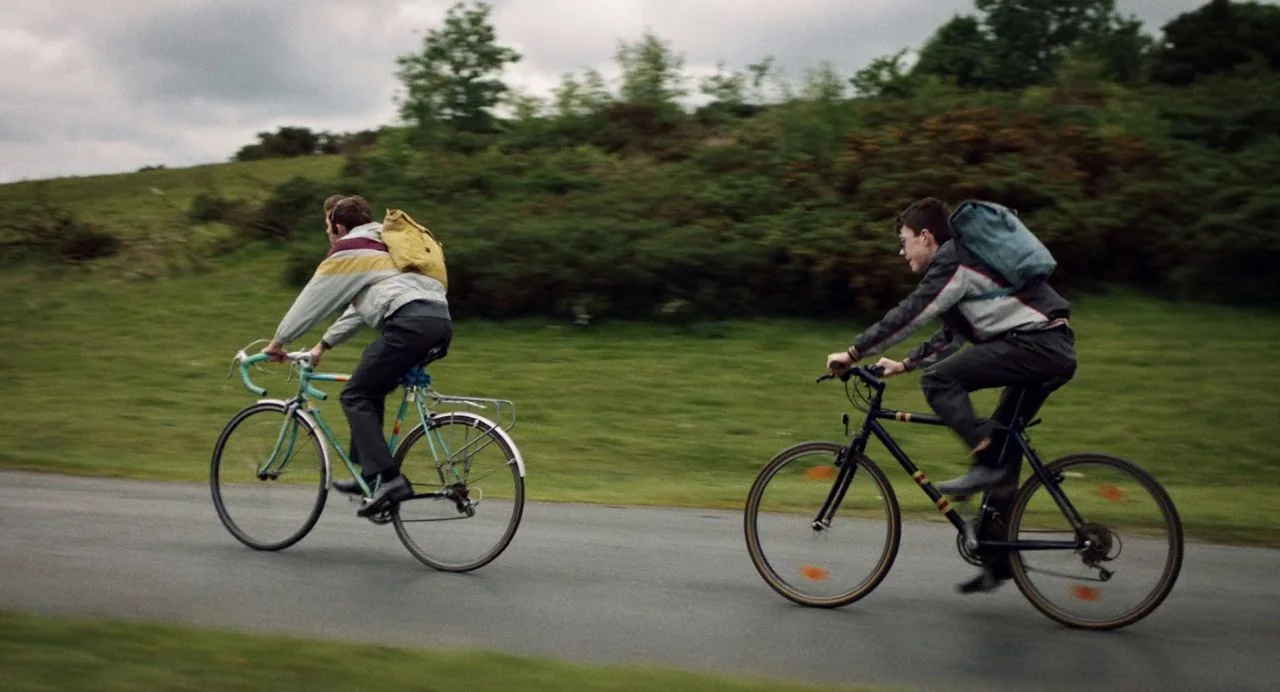 Dating Amber movie still: wide — A rural asphalt road cuts through a green, grassy landscape with rolling hills. Two teenage boys ride bicycles in…