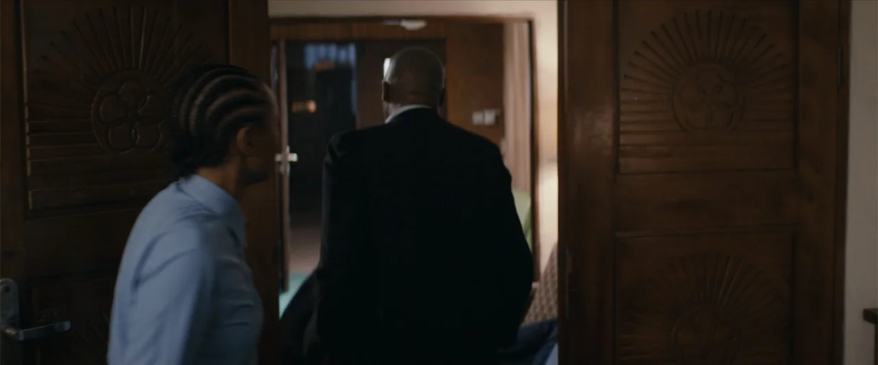 Ms. Kanyin (2025) movie still: medium — An interior doorway of a hotel suite features carved wooden panels with circular patterns. A man in a dark…