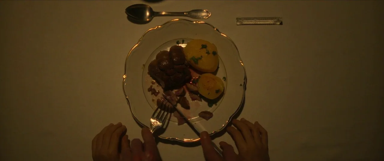 Earwig (2022) movie still: closeup — A white plate with roasted meat and potatoes sits on a dark table surface. Hands manipulate a silver fork and…