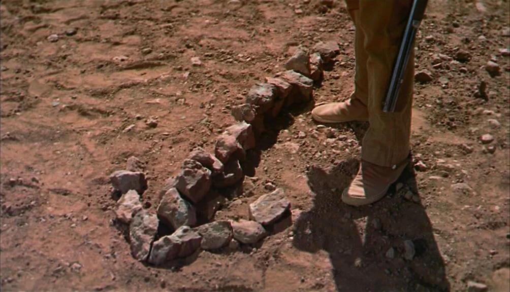 The Searchers movie still: closeup — Dry, rocky desert ground features a small pile of stones placed in the dirt. A person standing in tan trousers…