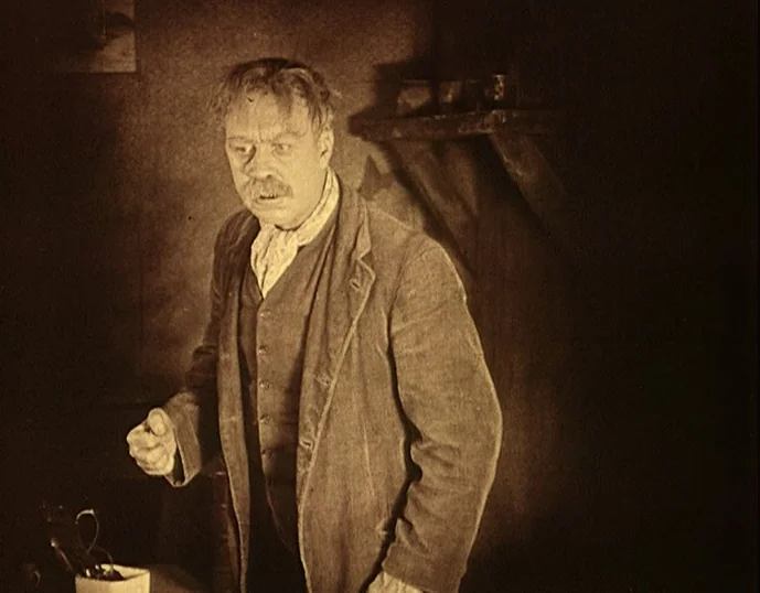 Broken Blossoms (1919) movie still: medium — A dimly lit room with bare walls and a wooden shelf mounted in shadow. A middle-aged man in a suit…